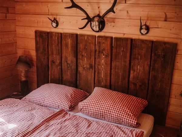 blockhaus jagdhuette schlafzimmer mit einem Doppelbett mit einem Braunen Holz Ramen und Hellen Holz Wänden Links daneben ein kleiner Nachttisch mit einer Lampe im Camping langenwald
