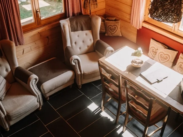 blockhaus landhaus, mit siecht von oben auf das Wohn- Esszimmer mit zwei Gemütlichen grauen Sesseln die am Fenster stehen. Rechts steht ein Esstisch mit einer Bank mit Kissen und zwei Stühlen. Mit Dunklem Fliesenboden und hellen Holz Wänden im Camping langenwald